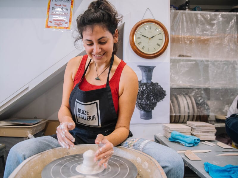 A woman in a black printed apron sitting at a potters wheel is starting to throw some clay
